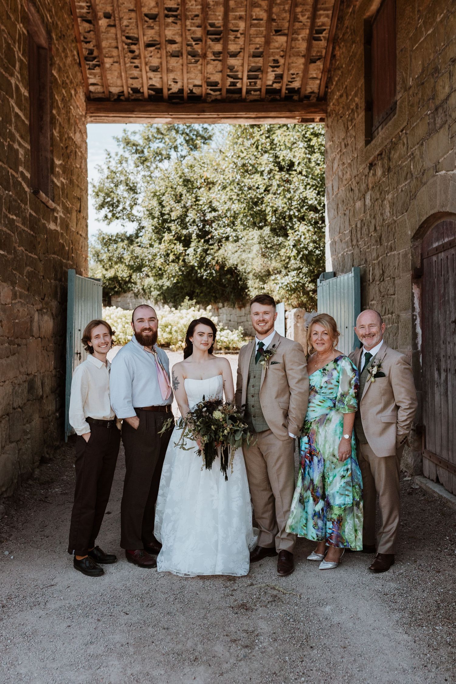Wedding party poses in a rustic stone barn doorway wearing tan suits and formal attire for group portraits at The Courtyard Wedding Venue