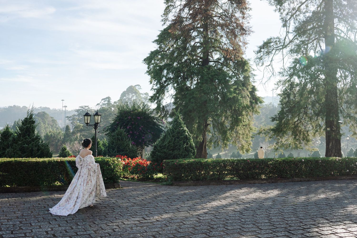 Bride in floral gown twirling near a lamp post in a scenic park with tall trees and blue sky.