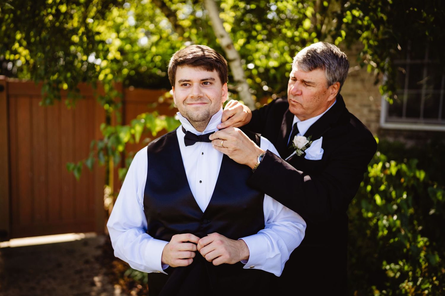 Two people in formal attire share a playful moment outdoors with one adjusting the other's collar in sunlit setting.