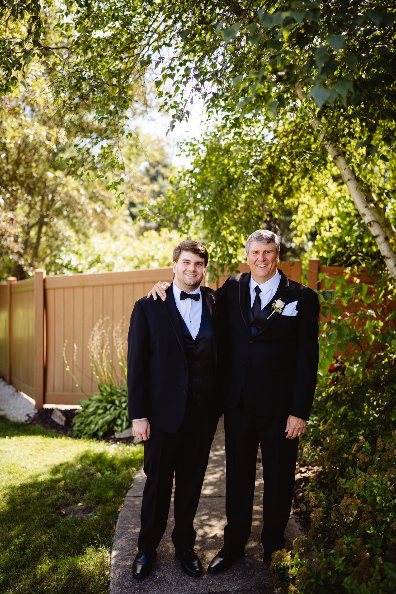 Two people wearing black formal attire stand together in a garden setting with trees and a wooden fence in the background.