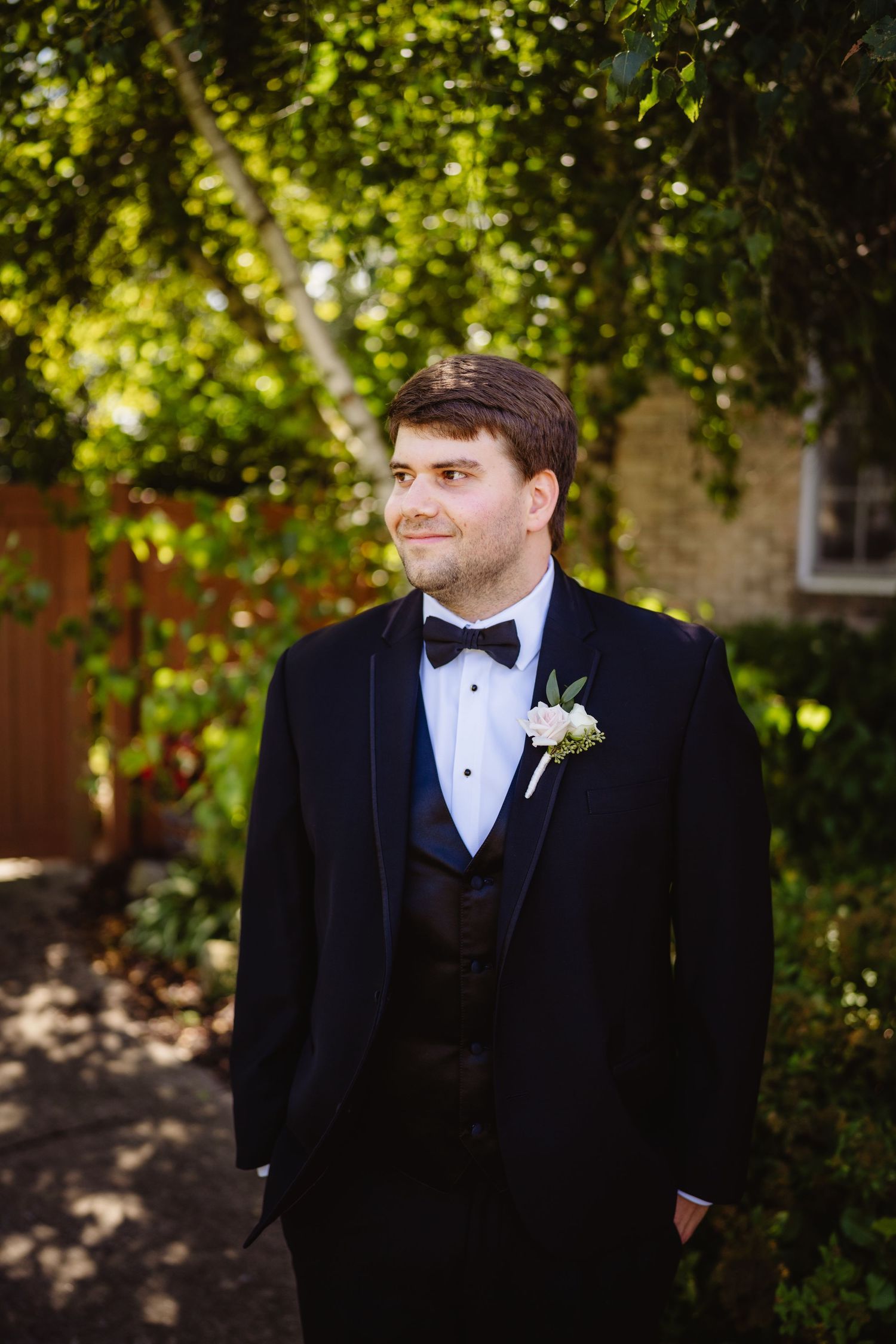 Groom in classic black tuxedo poses outdoors against lush green garden background.