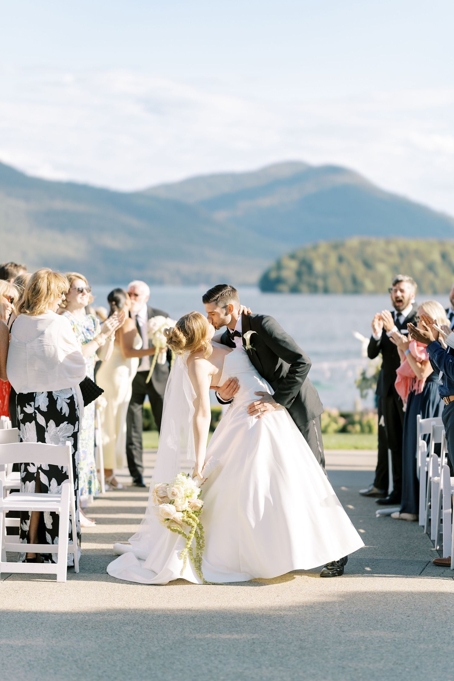 Wedding party arranged in a dip kiss pose along a lakefront ceremony site with mountain views in the background.
