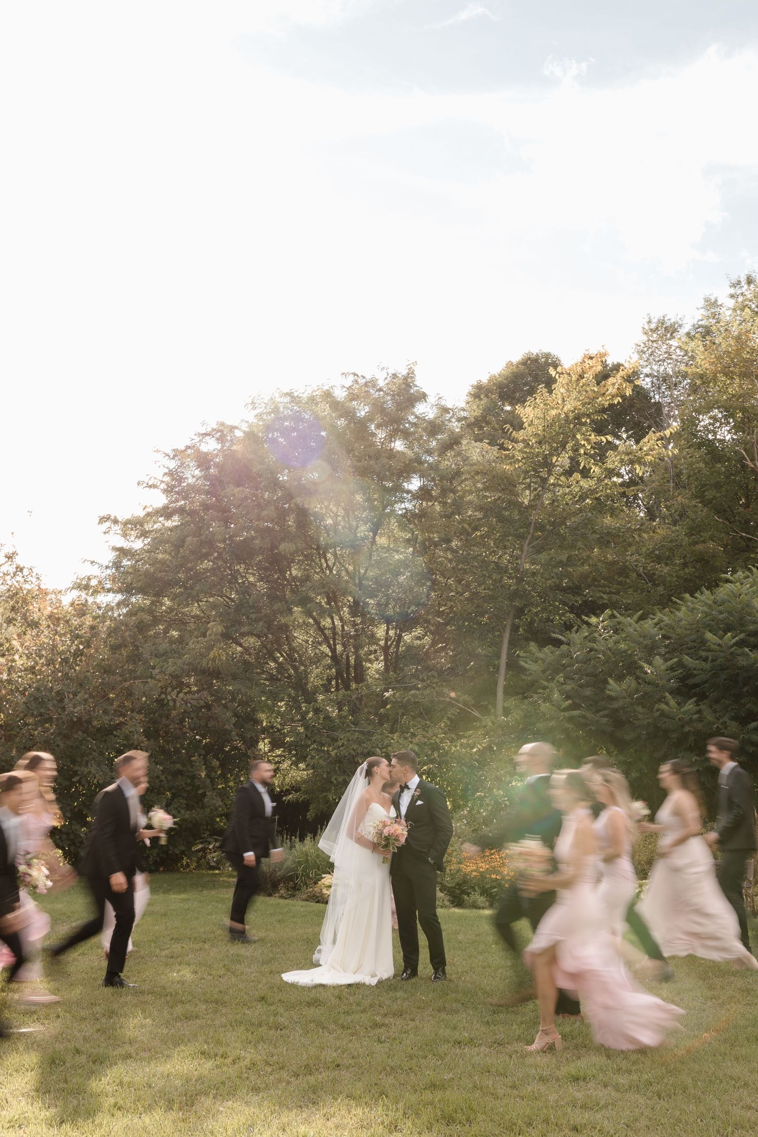 Artistic wedding photography with couple kissing and wedding party running around them, captured with them being blurry. Fun and creative.