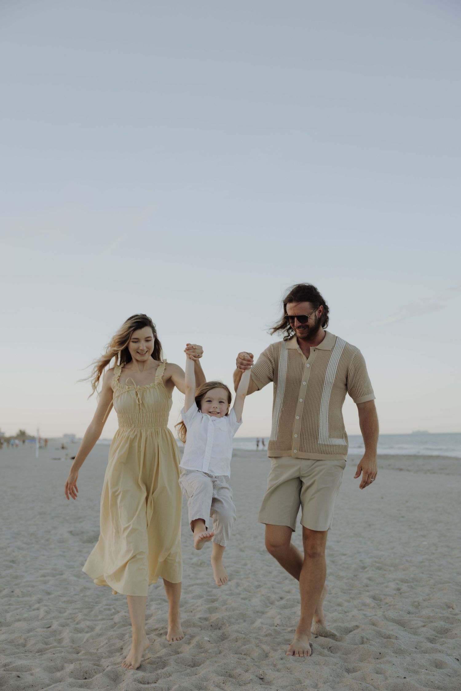 A family enjoys a playful moment together on the beach at sunset, dressed in neutral tones.