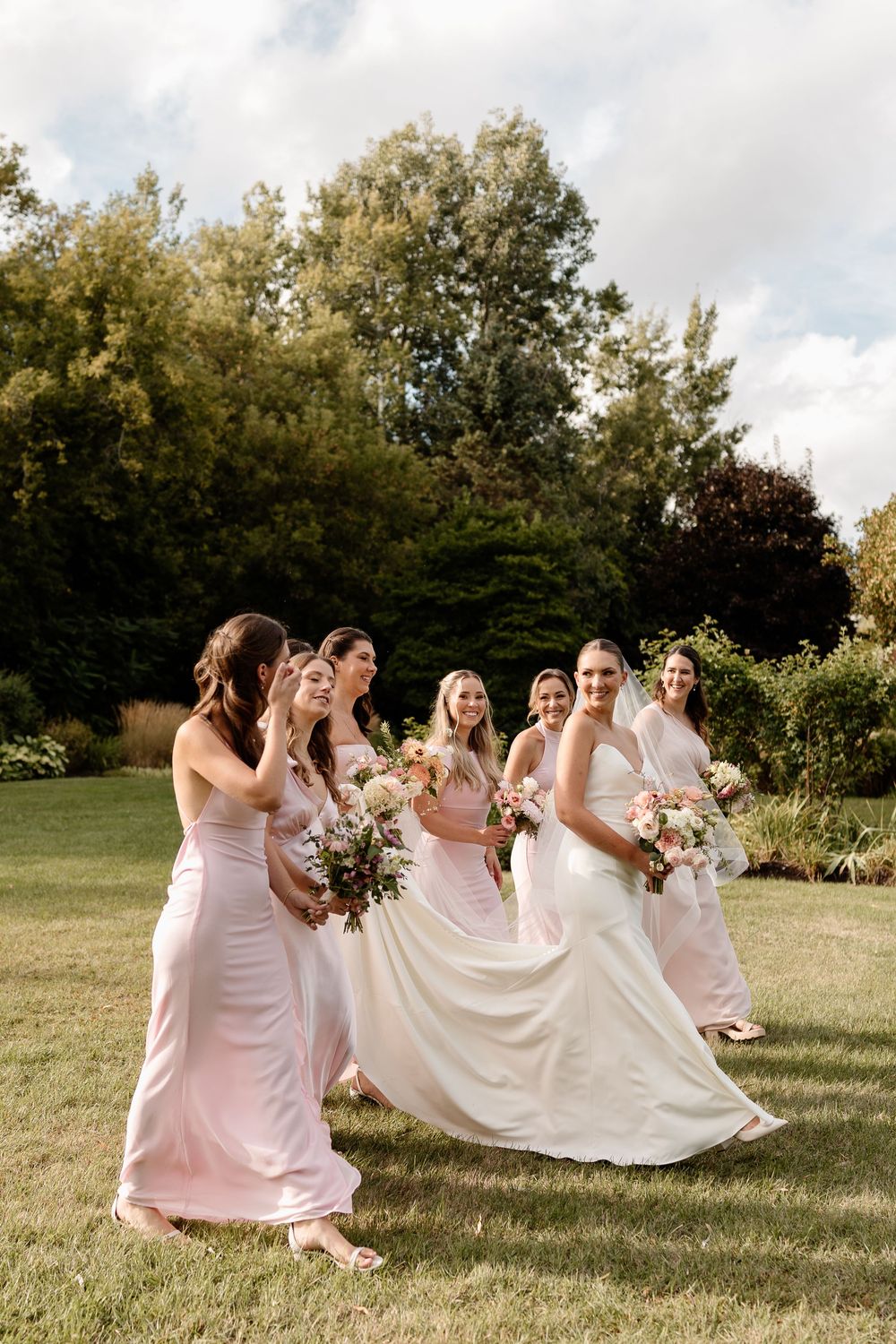 Bridal party in pink dresses laughs and plays with the bride's wedding dress train in a sunny outdoor field.