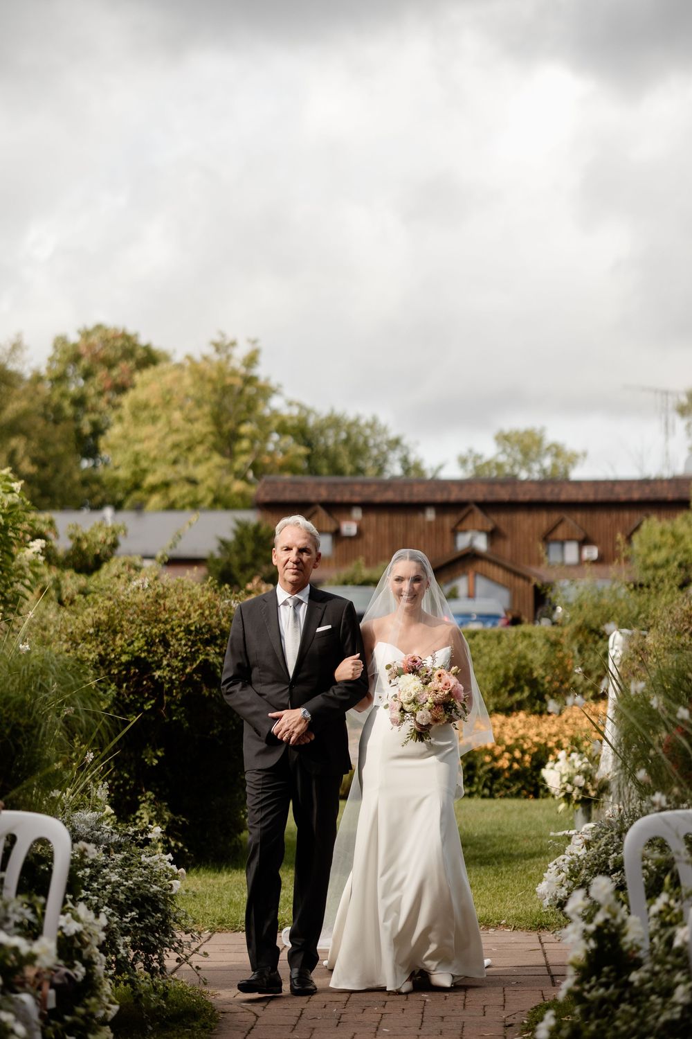 Wedding ceremony scene in a garden setting with guests seated and floral decorations on a cloudy day.