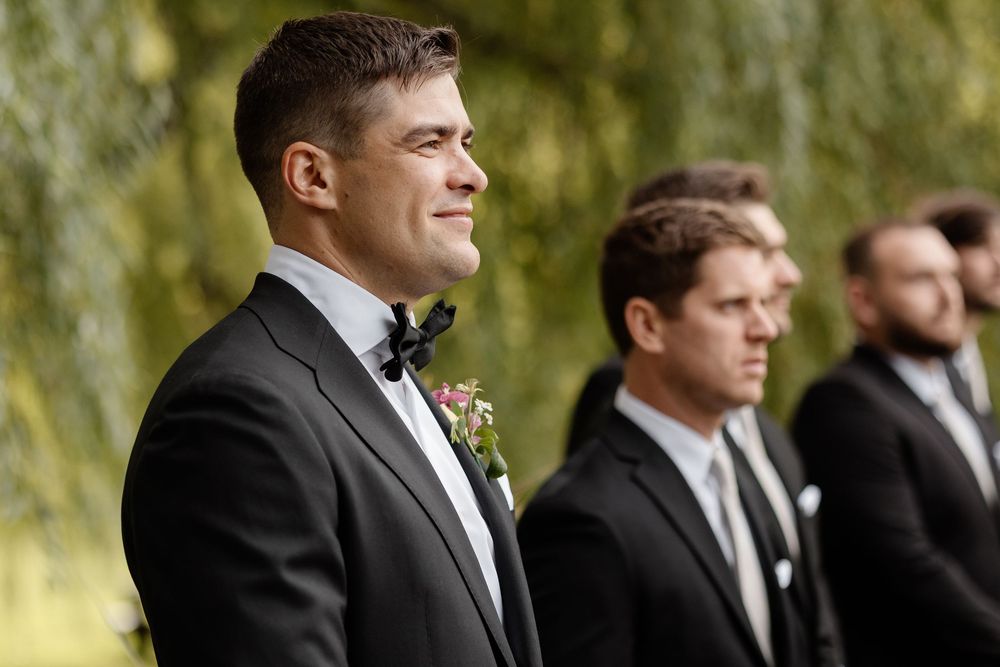 A wedding party in formal black suits stands in a row outdoors with willow trees in the background.