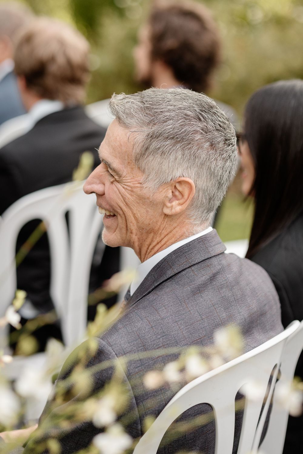 A person in a gray suit smiles while seated in a white chair at an outdoor event.