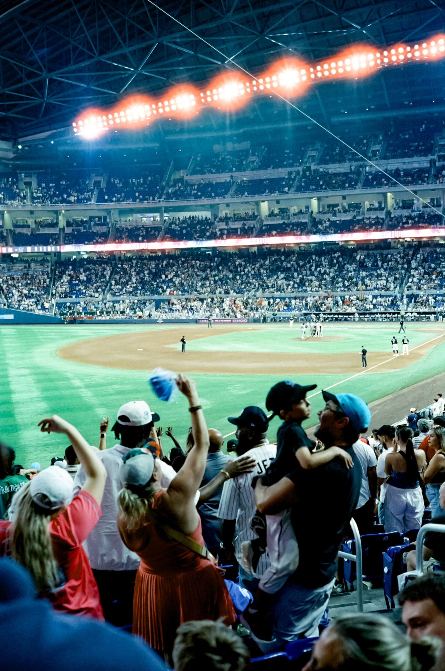 Excited baseball fans cheer from the stands at a night game in a large stadium with bright overhead lights illuminating the field.