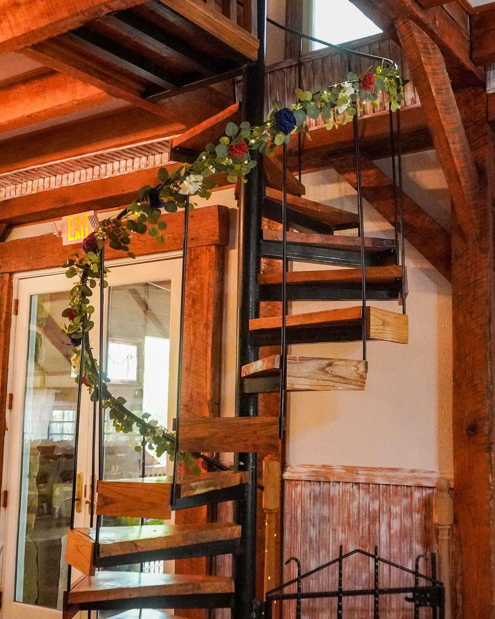Interior view of rustic restaurant with exposed wooden beams, spiral staircase, and hanging plants creating warm ambiance.