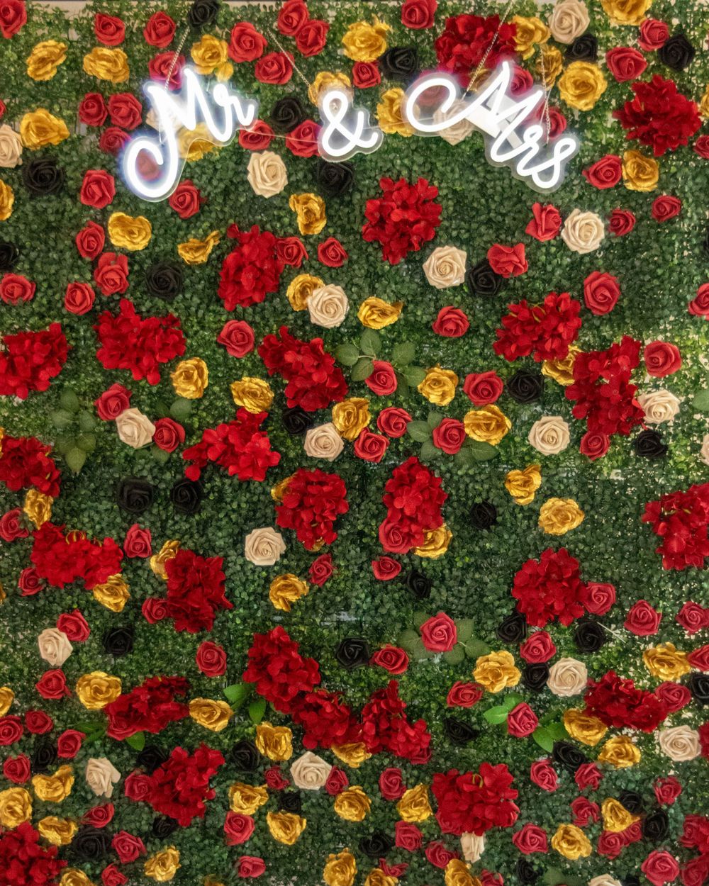 Floral wall backdrop with neon 'Mr & Mrs' sign amid red, yellow and cream roses against lush greenery.