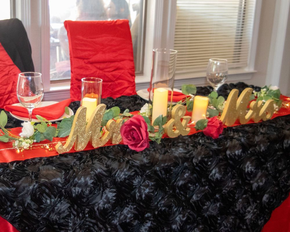 A wedding head table features gold 'Mr & Mrs' lettering, red roses, and champagne glasses on a black textured tablecloth.