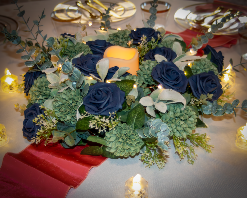 A romantic table setting features navy blue roses, greenery, and twinkling lights along a red runner at a wedding reception.
