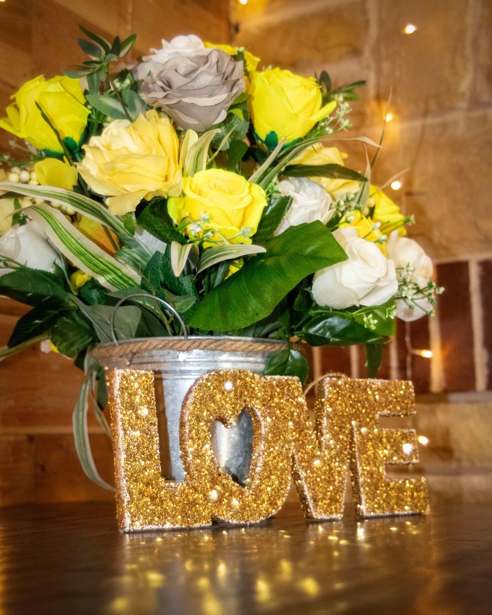 Gold glittery 'LOVE' letters arranged with yellow and white roses in silver vases on a wooden table at a wedding reception.