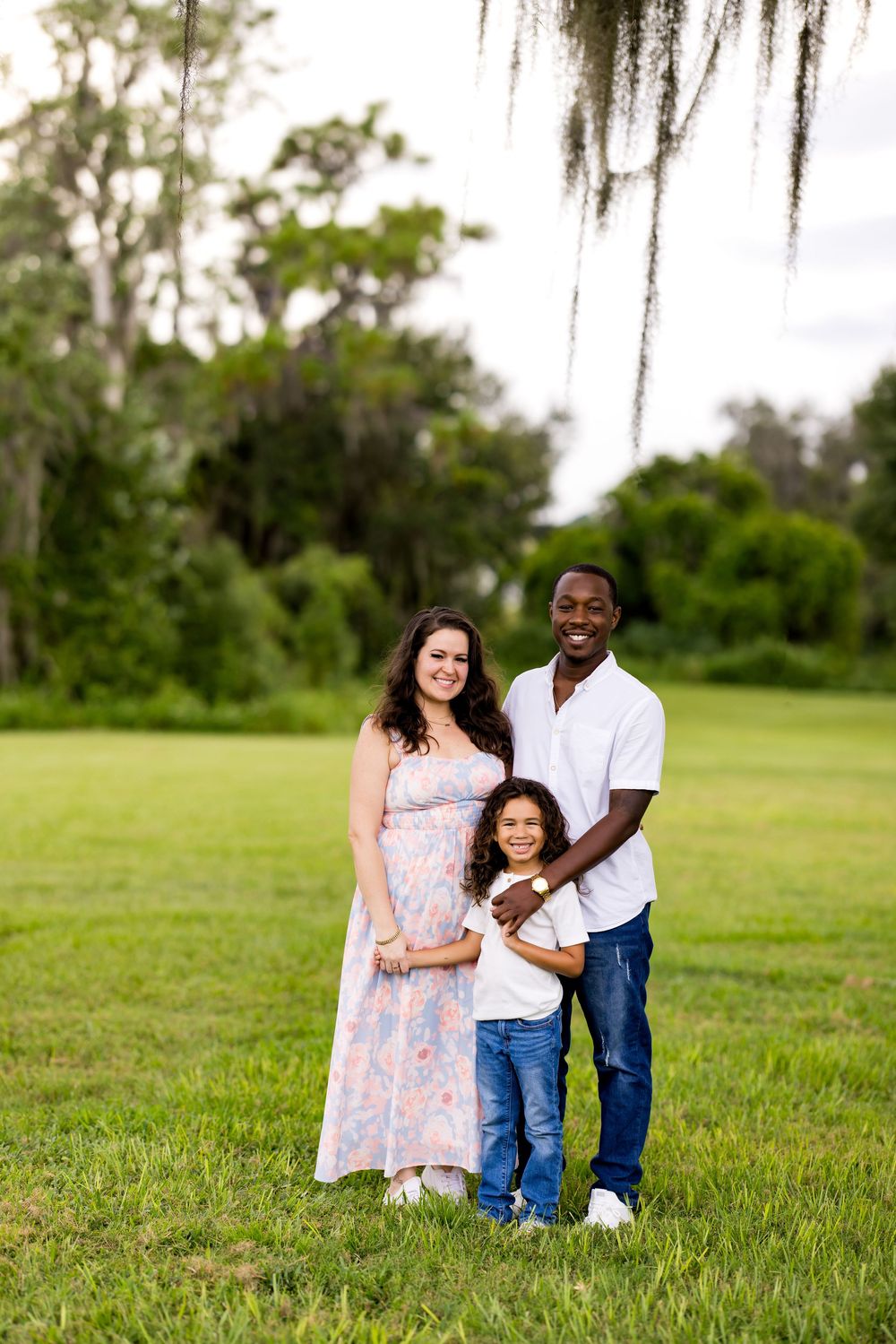 A loving family enjoys a tender moment together while sitting on lush green grass beneath Spanish moss trees.