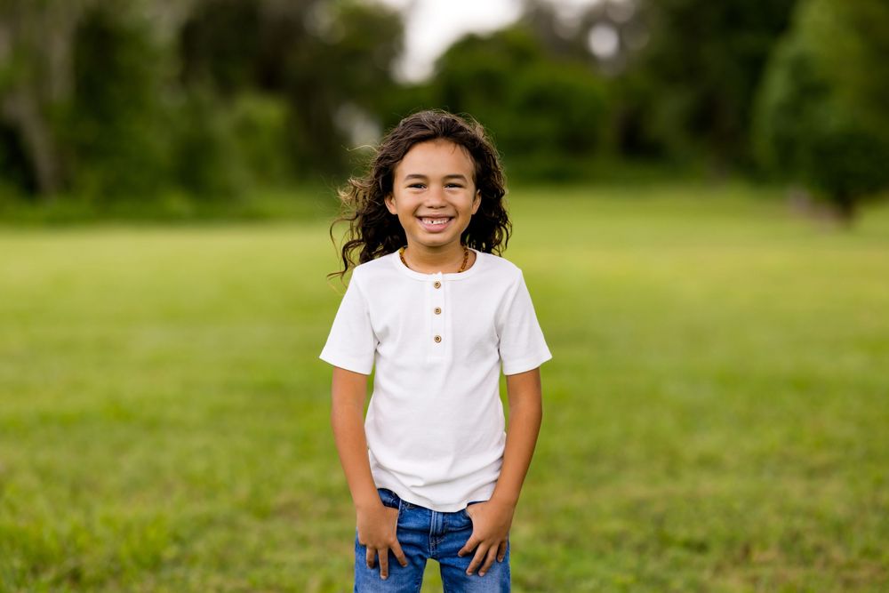 A happy person in a white t-shirt and blue pants sits on grass in a lush outdoor setting during daytime.