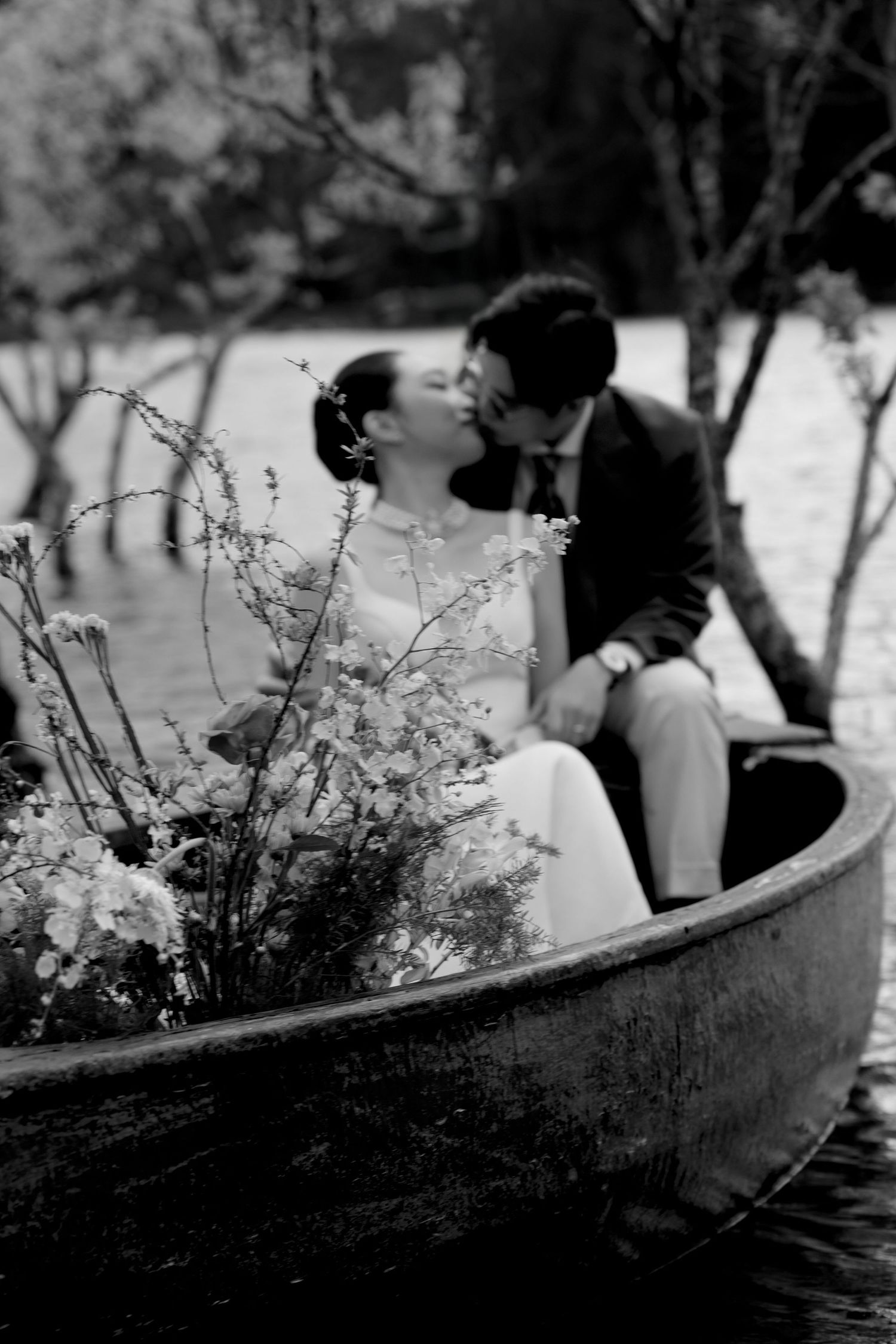Black and white romantic photo of a couple kissing in a flower-filled wooden boat surrounded by trees.