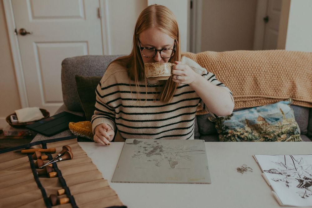 An artist works at a desk with papers and art supplies while drinking from a mug. Whitney Popp Arizona photographer