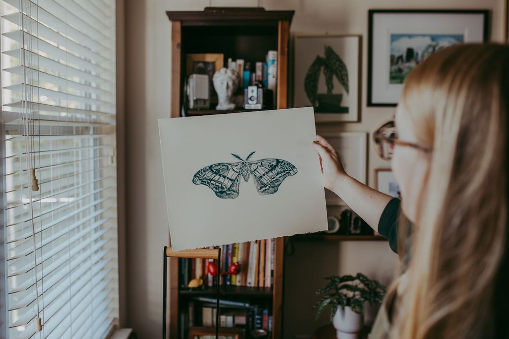 A person sits by a window with venetian blinds in a cozy home office setting with framed artwork on the wall looking a linocut image.