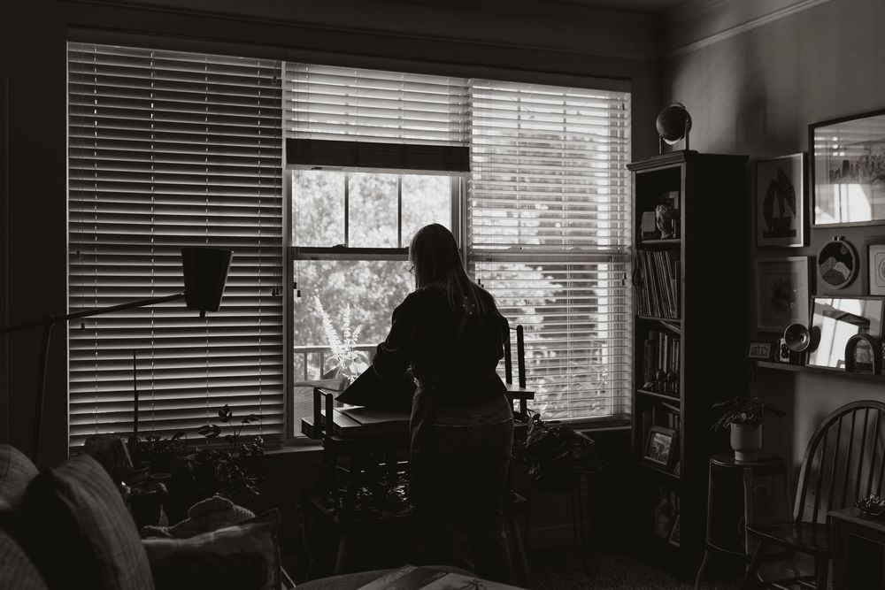 A black and white photo showing a dark silhouette against a bright window with venetian blinds in an office setting. Whitney Popp, Arizona.