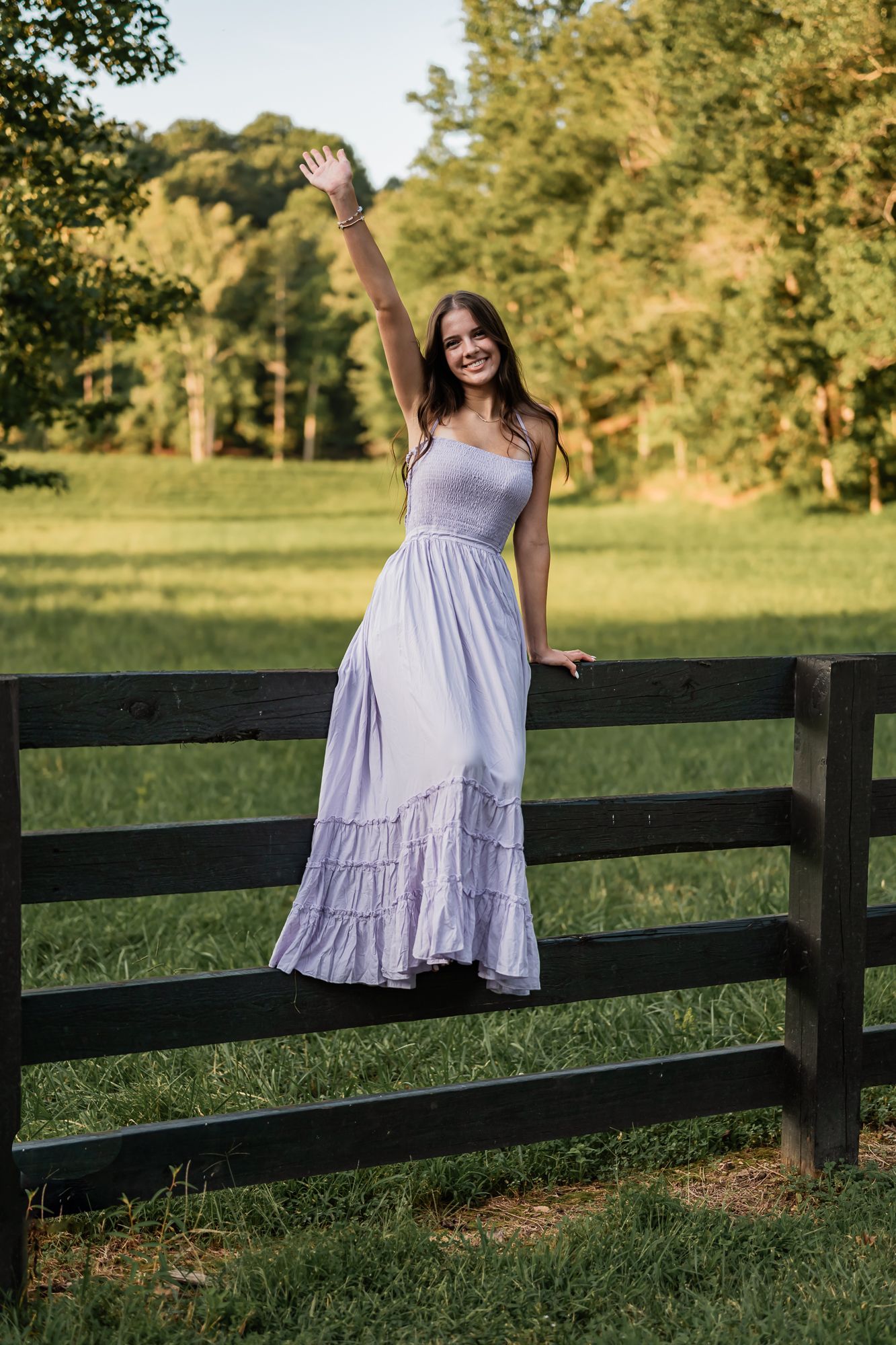 A person in a flowing lavender dress poses against a black wooden fence in a sunlit country field at golden hour.