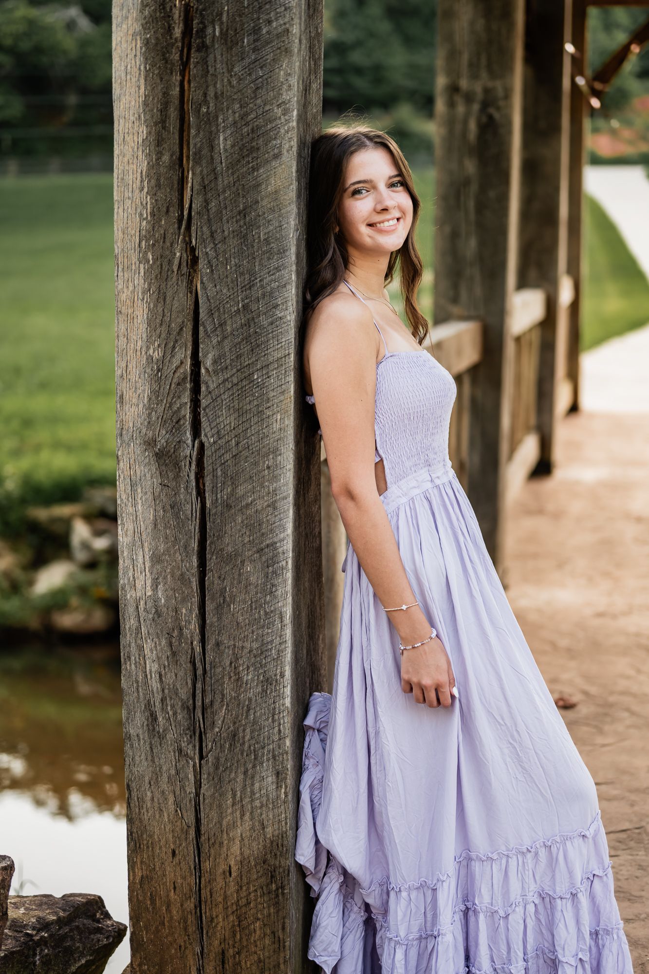A person in a flowing lilac summer dress poses gracefully against rustic wooden posts in an outdoor setting at sunset.