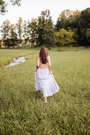 A figure in a flowing white dress twirls through a sunlit meadow with trees in the background.