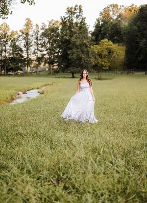 A white lace wedding dress flows in a serene meadow with a small stream and treeline in the background.