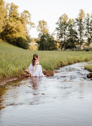 A person sits barefoot in a flowing stream surrounded by green grass and trees on a sunny summer afternoon.