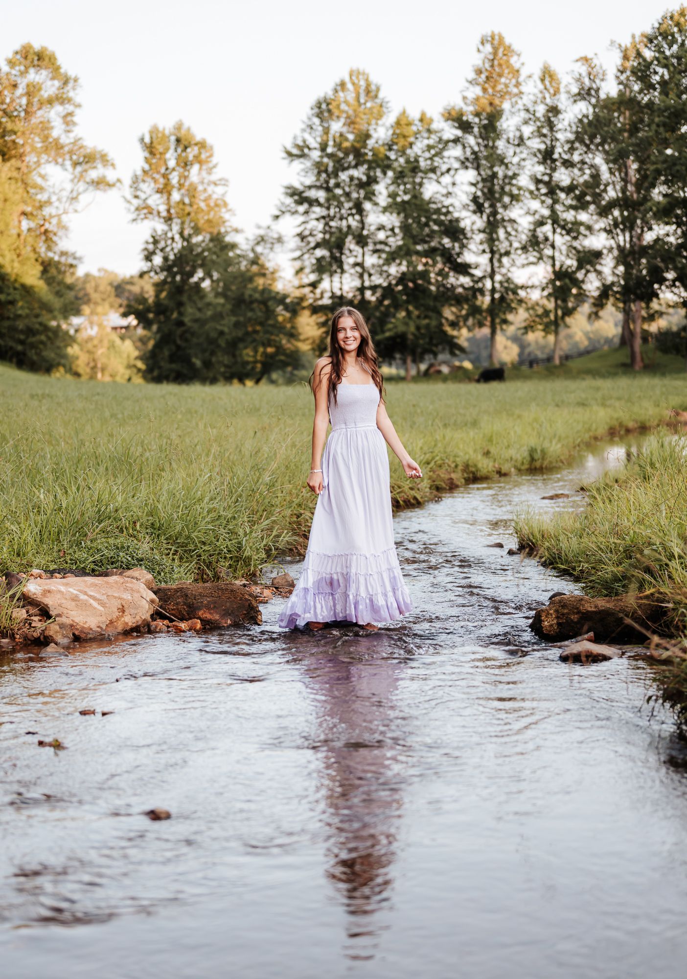 A serene white dress stands ethereally in a flowing stream surrounded by golden hour sunlight and lush greenery.