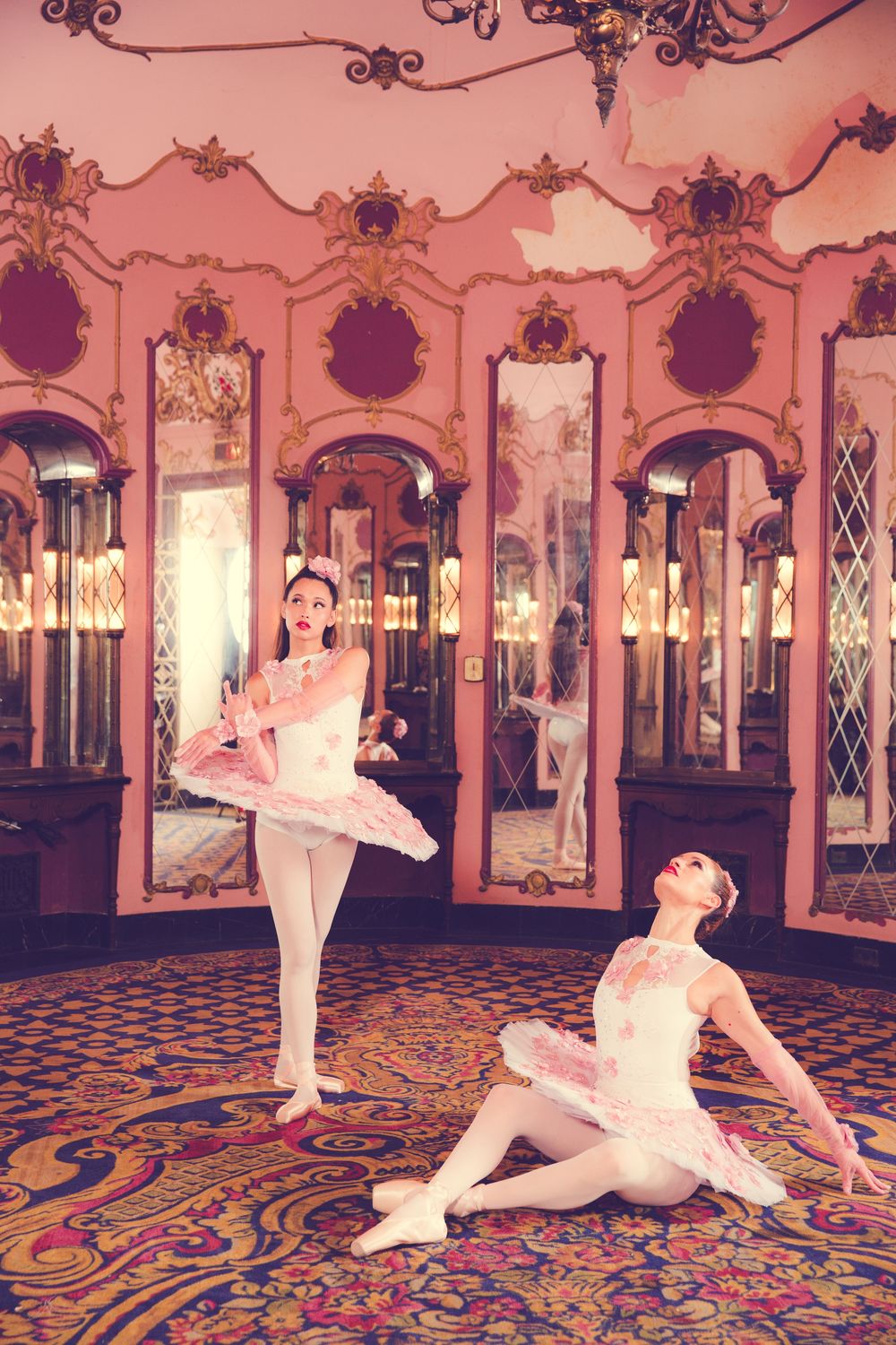 Two ballet dancers in white tutus strike graceful poses in an ornate mirrored ballroom with patterned carpet.