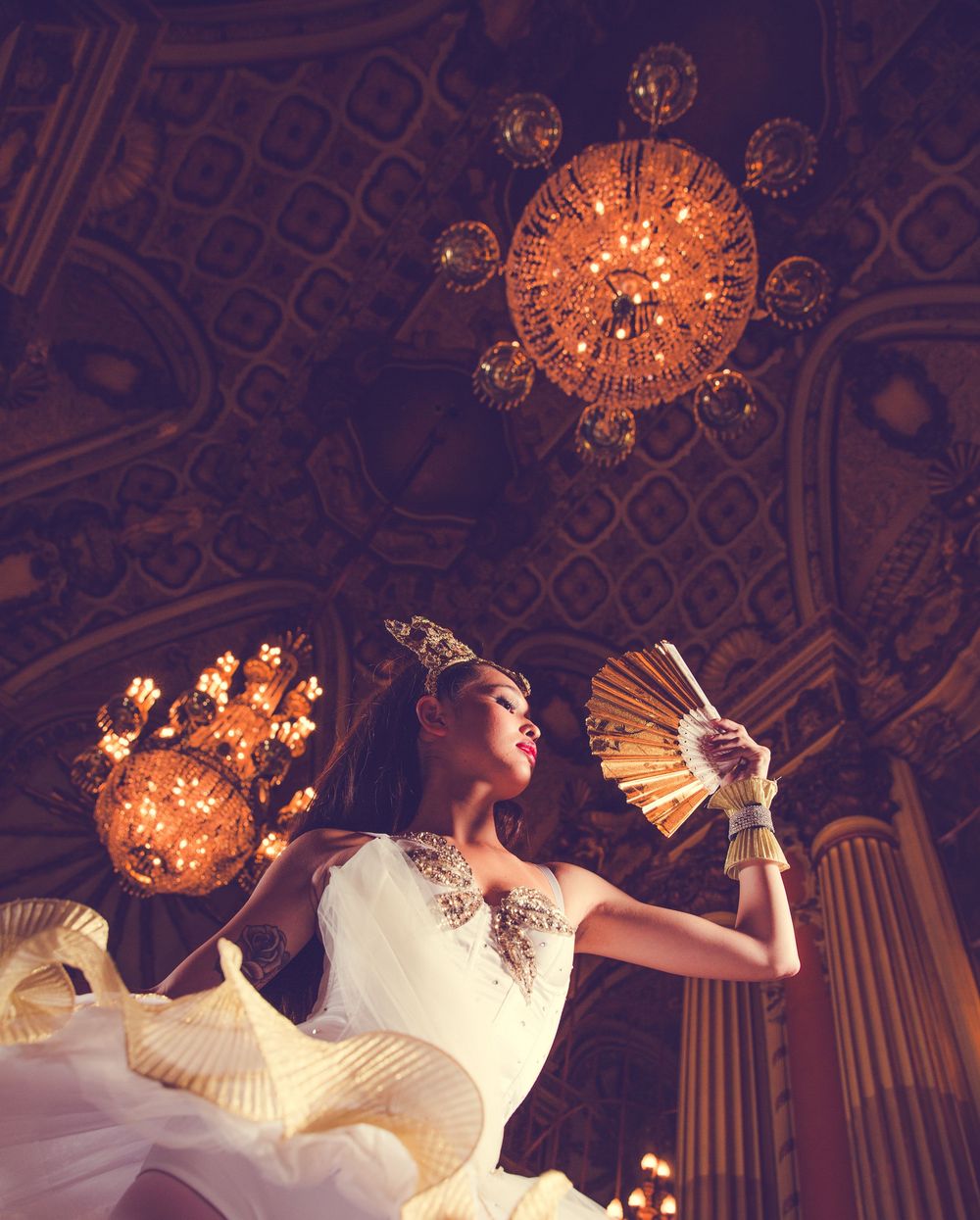 Dancer in white dress poses with fan under ornate crystal chandeliers in grand ballroom.