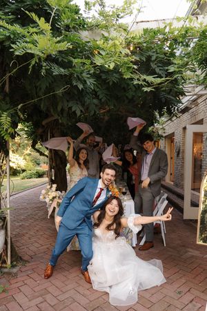 Wedding party dances and celebrates with energetic moves on an outdoor brick patio beneath leafy trees.