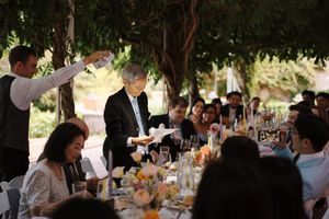 Wedding reception guests gathered at long tables under trees for an outdoor celebration.