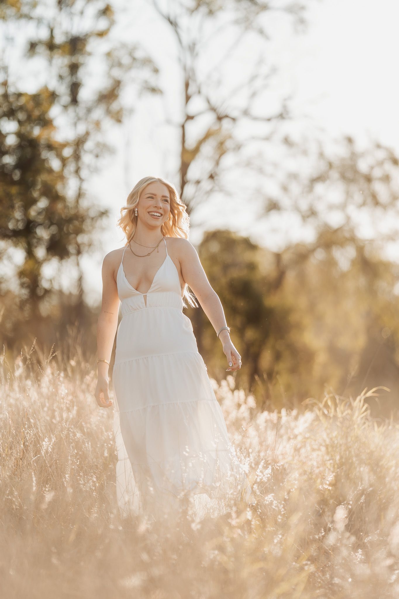 A person in a flowing white dress stands in a sun-drenched field of tall grass during golden hour.