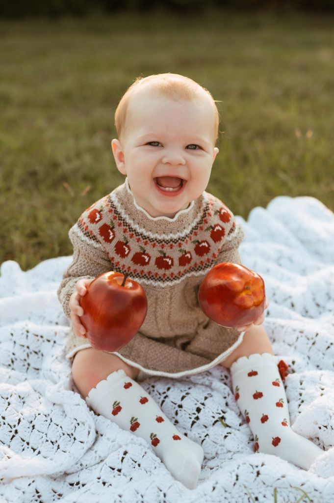 Baby plays with apples while sitting on lace blanket wearing tan knit sweater with apple pattern.