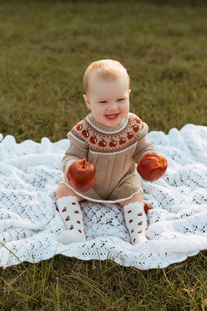Smiling baby wearing festive sweater plays with small pumpkins on white lace blanket outdoors.