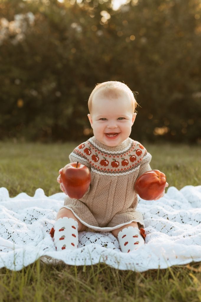 Smiling infant in apple-patterned sweater plays with red apples on delicate white lace during autumn photoshoot.
