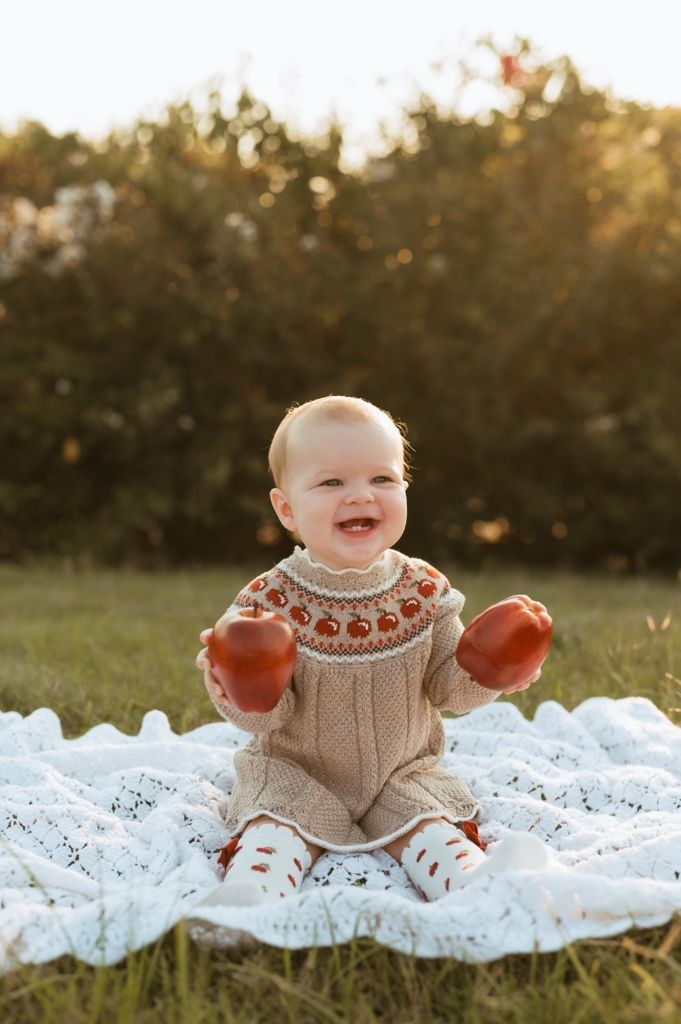 Happy baby in cozy knit sweater poses with pumpkins on white lace blanket during golden hour.
