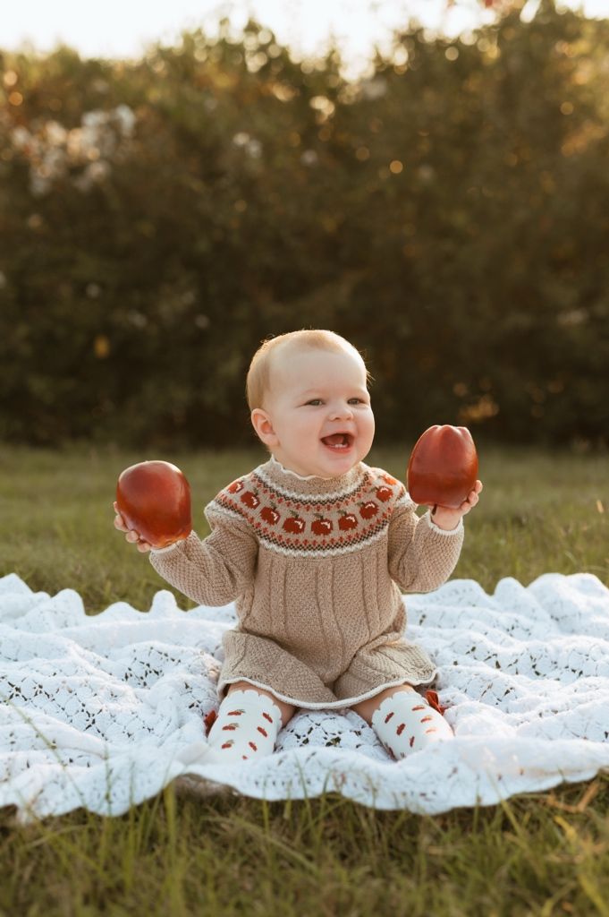 Joyful baby wearing beige knit sweater holds red apples while sitting on white lace outdoors.
