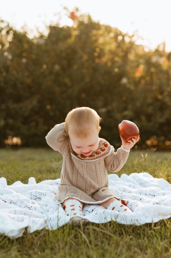 Baby in cozy apple-patterned sweater plays with fruit on white lace blanket during sunset.