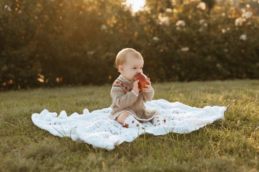 Baby in white dress sitting on blanket against golden sunset backdrop in grassy field.