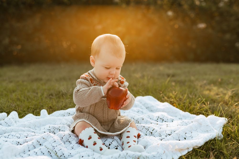 Baby in a beige sweater plays with a red ball while sitting on a blue blanket outdoors at sunset.
