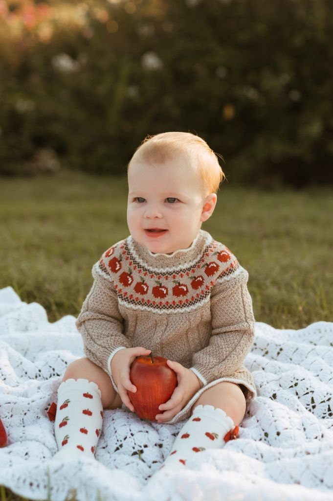 Baby wearing cream-colored sweater with pumpkin design sits on white blanket holding orange pumpkin in fall setting.