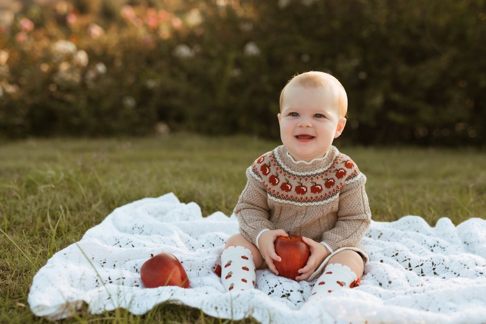 A small infant wearing a beige sweater with apple patterns plays on a white lace blanket in an outdoor autumn setting.