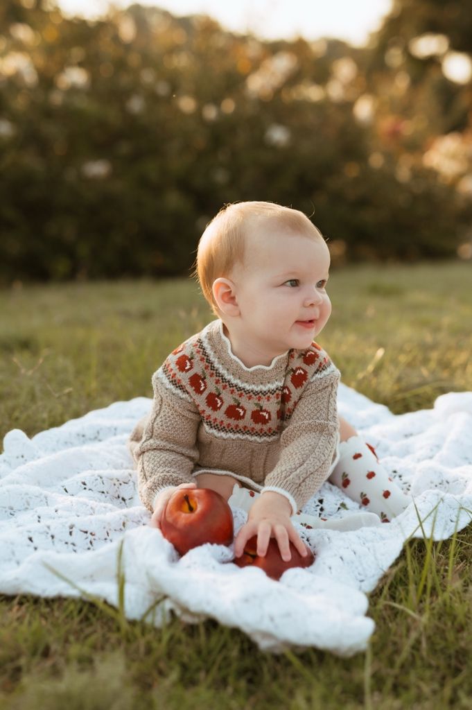 Baby in knit sweater with red pattern lies on white lace blanket on grass during autumn photoshoot.