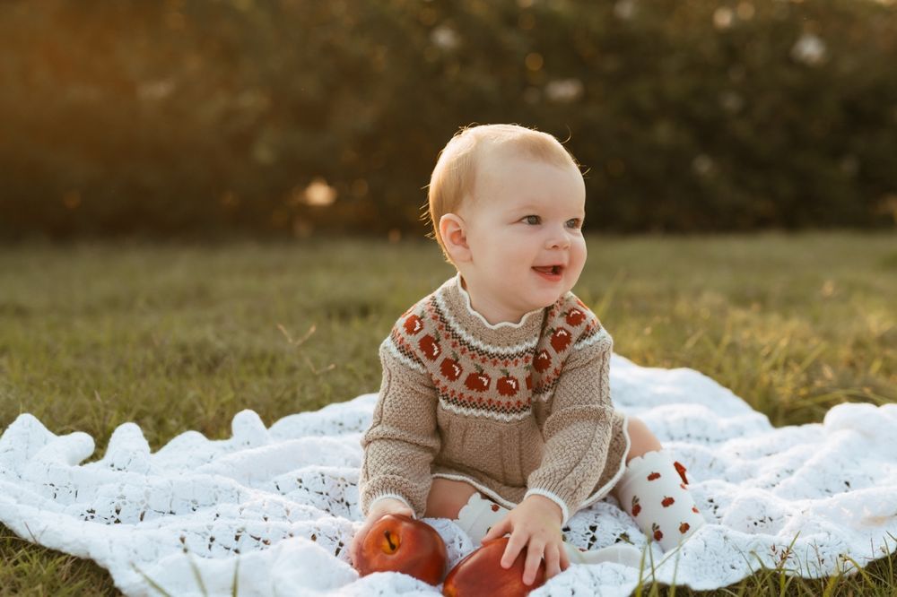 Baby in knit sweater holds red apples while sitting on white lace blanket outdoors during golden hour.