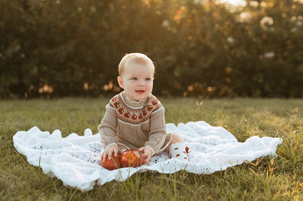 Baby sits on white blanket outdoors wearing knit sweater with autumn pumpkins during sunset photoshoot.