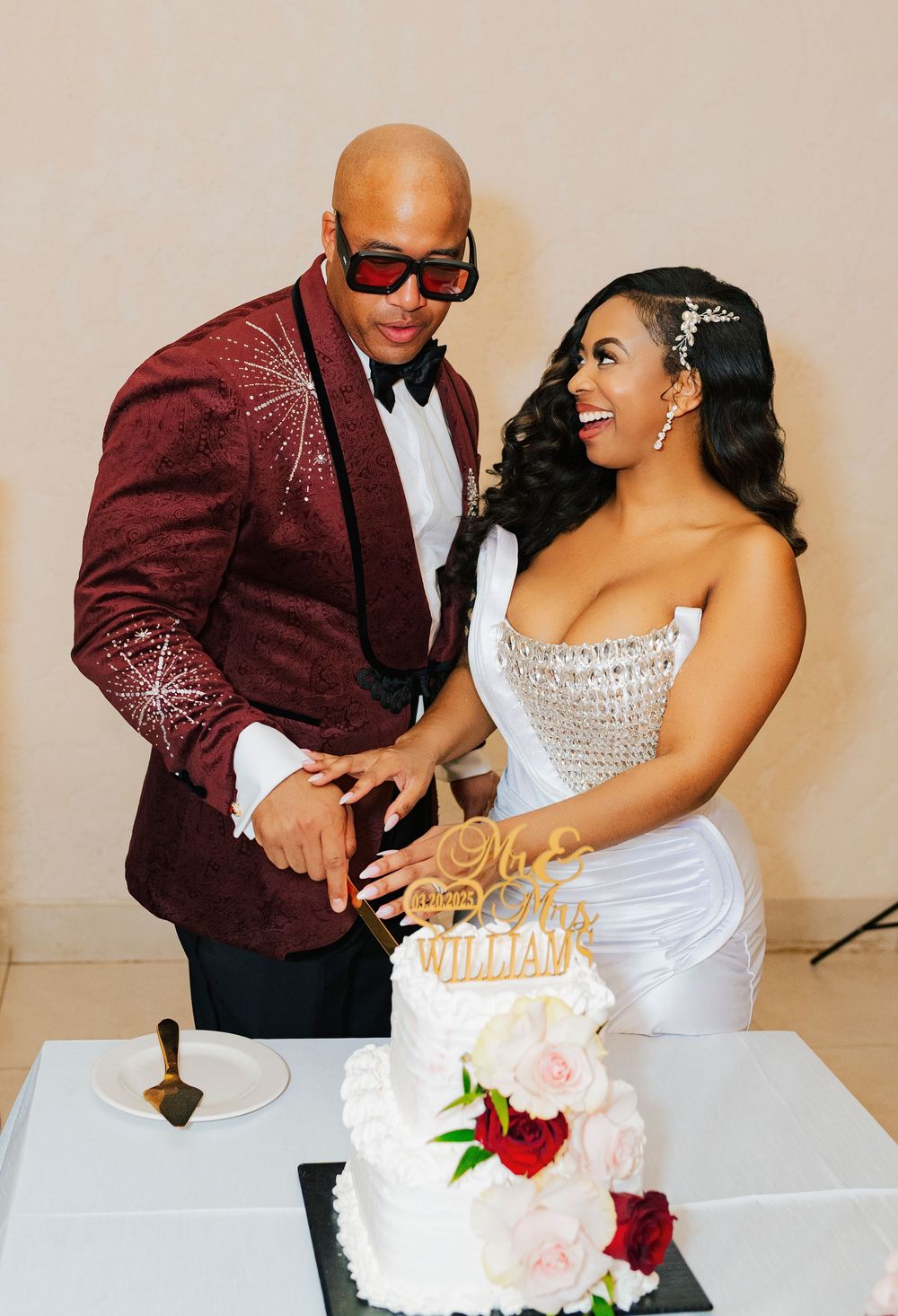 A couple in formal attire cuts their elegant white and red wedding cake at their reception celebration.