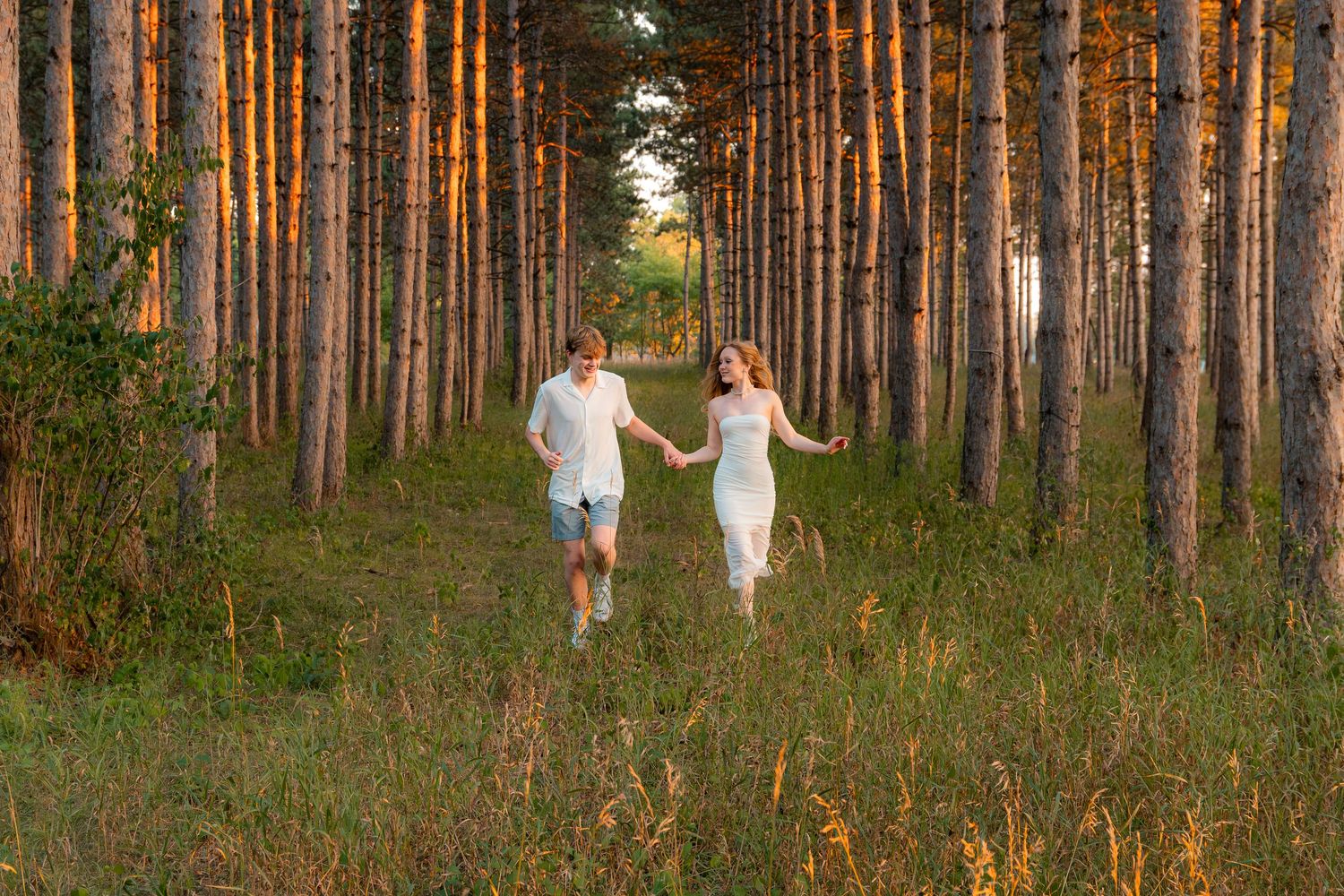 Couple holding hands running through a sunlit pine forest path at golden hour in white summer outfits.